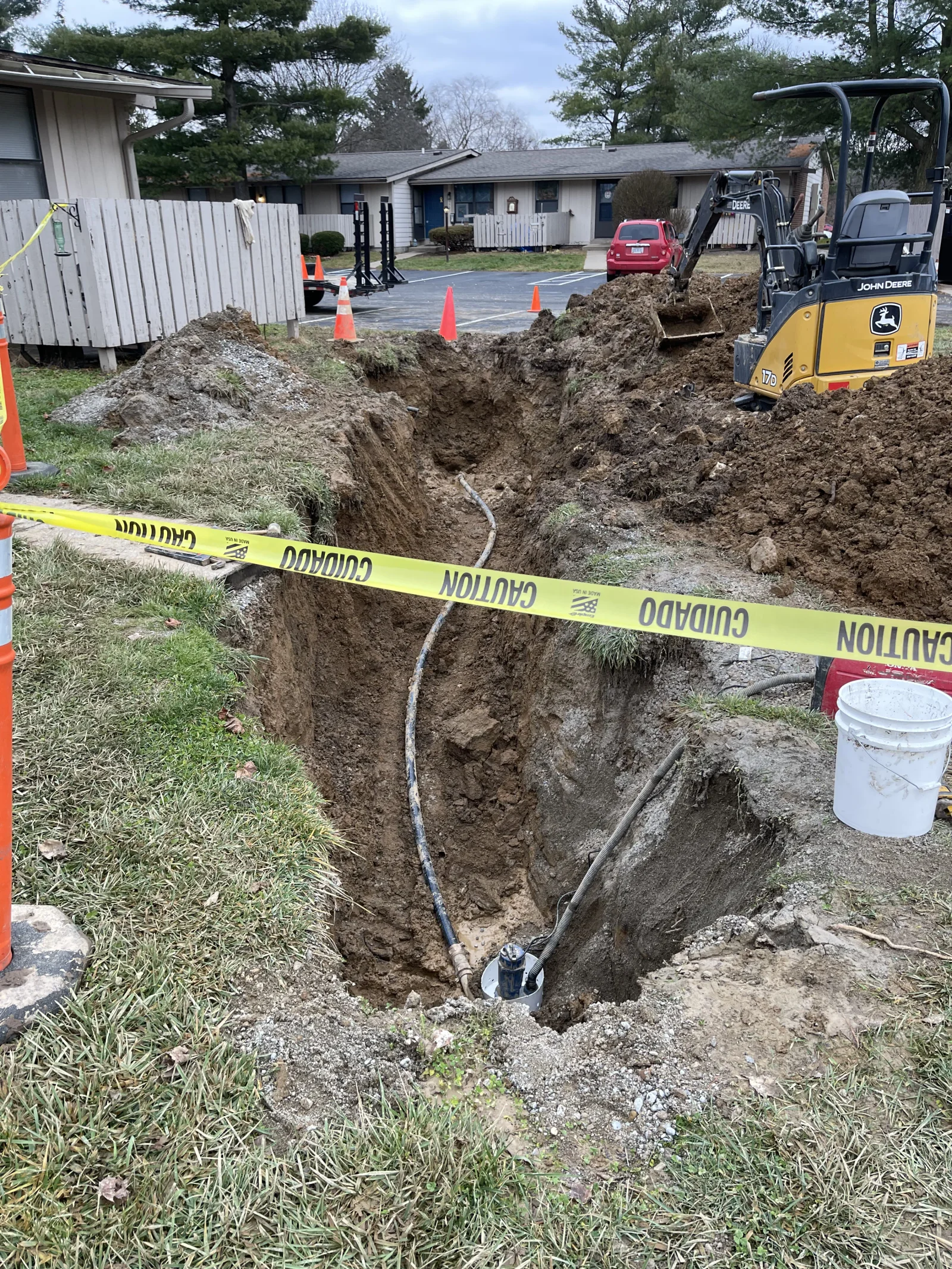Excavated trench exposing underground sewer pipe with mini excavator and caution tape at residential property