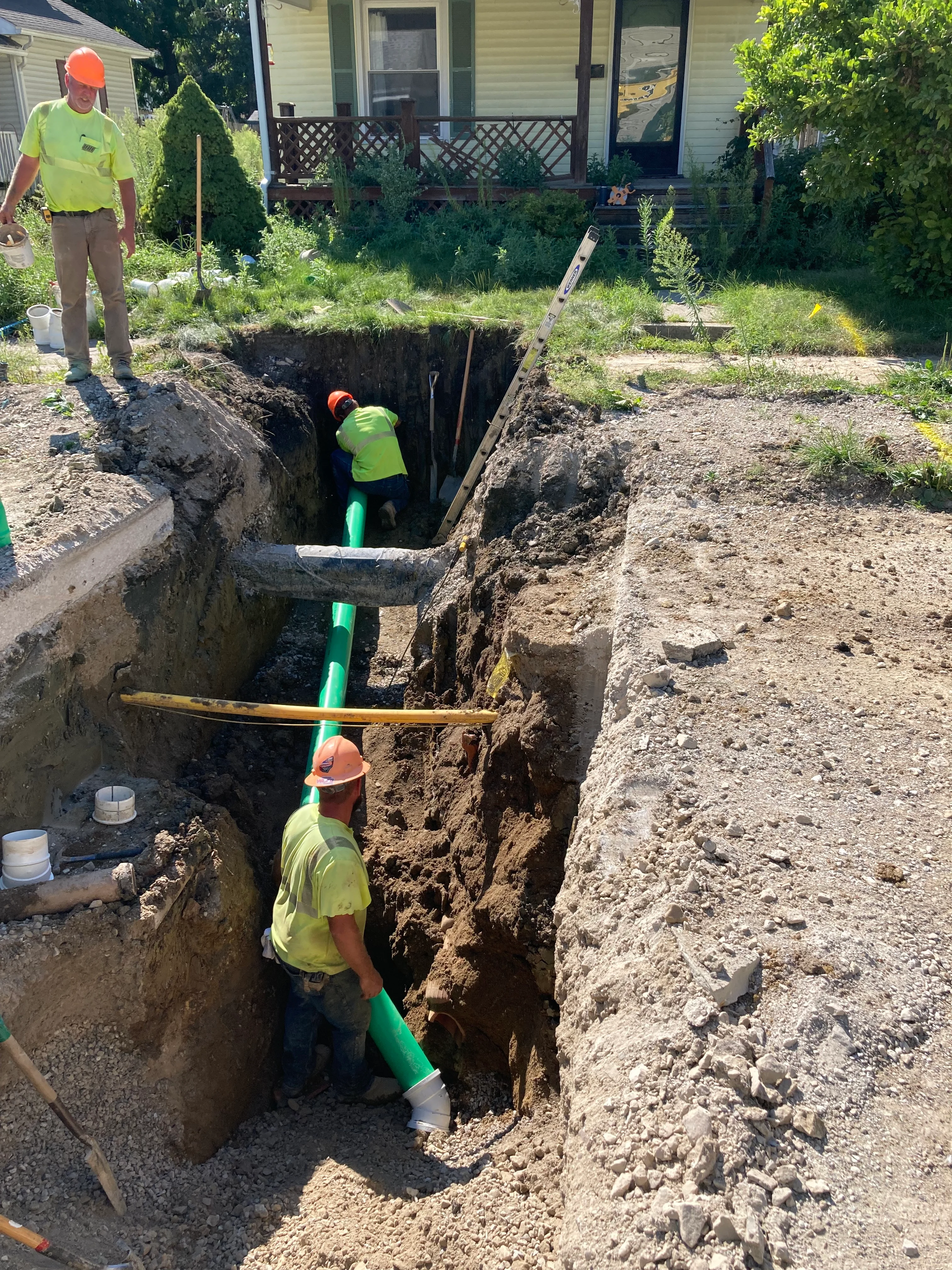 Crew installing new green PVC sewer pipe in deep residential trench excavation
