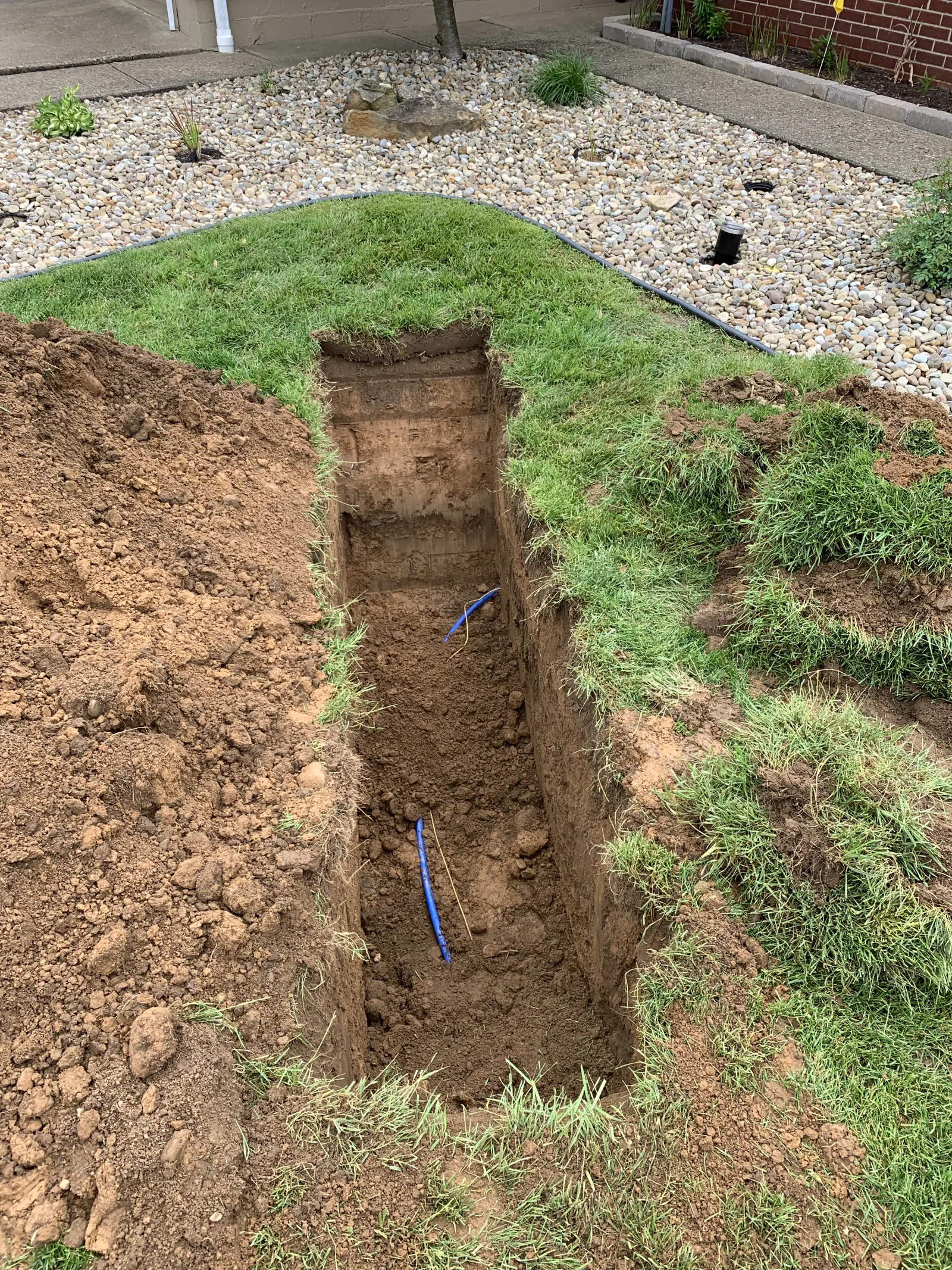 Deep trench with blue PVC water line pipe installed in soil, surrounded by excavated earth and grass