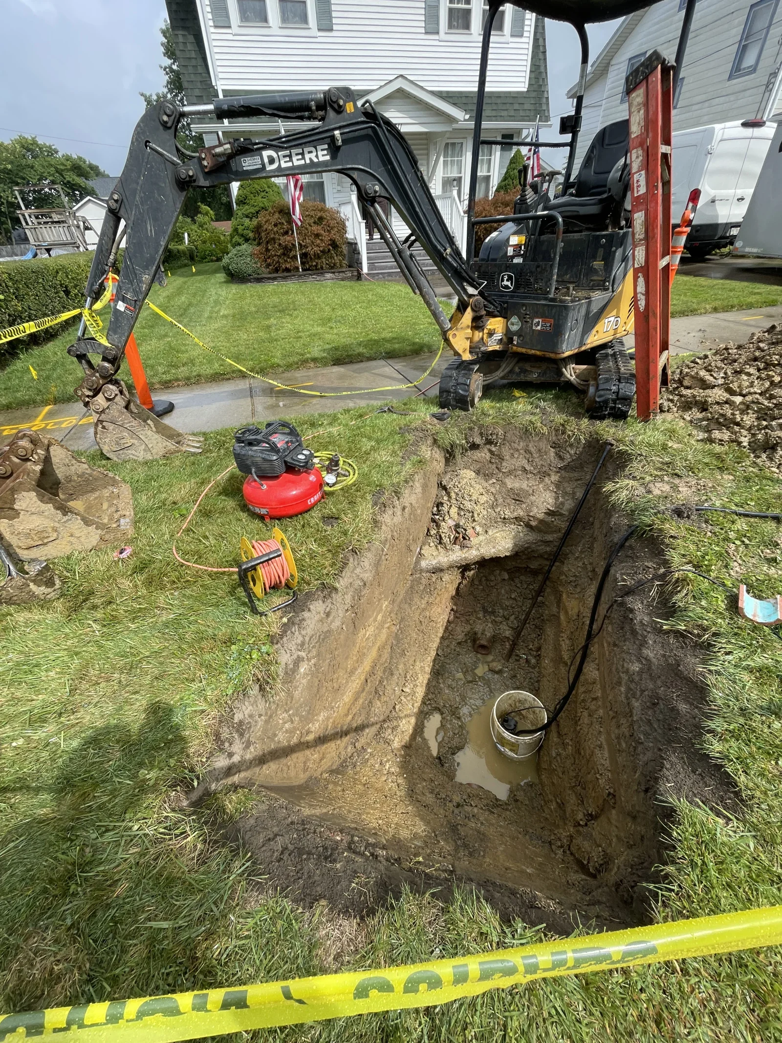Compact excavator digging deep residential drainage trench with safety equipment and air compressor on site