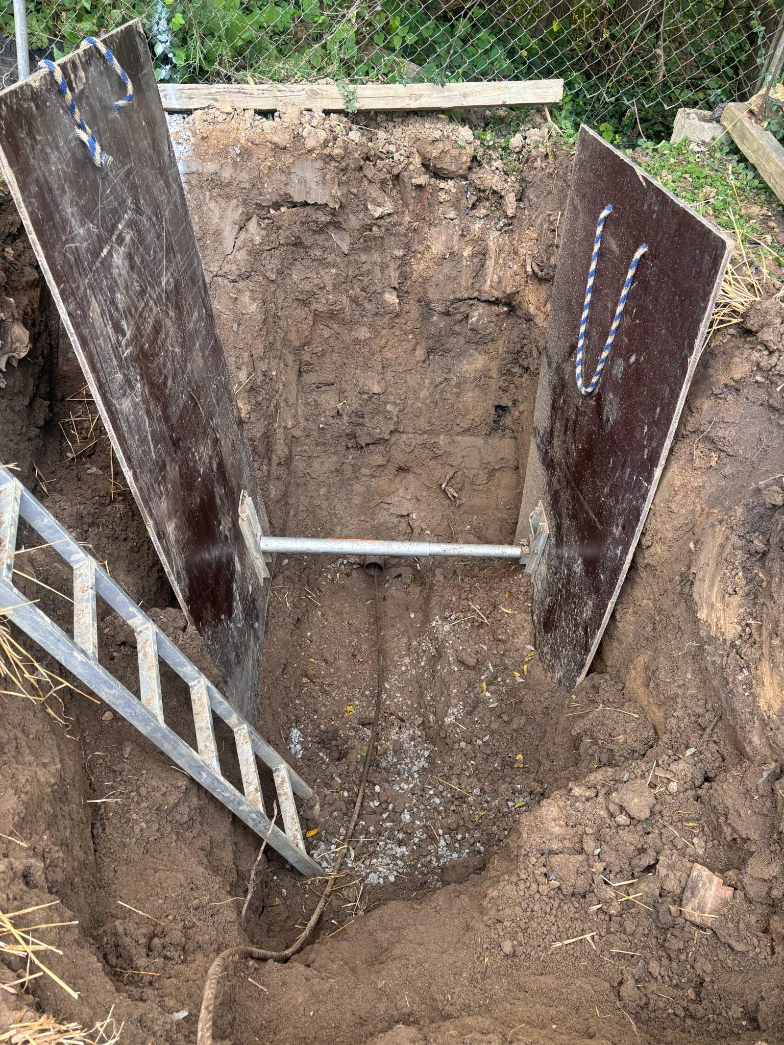 Excavated trench with plywood shoring, wooden bracing, and utility line markings for drainage installation