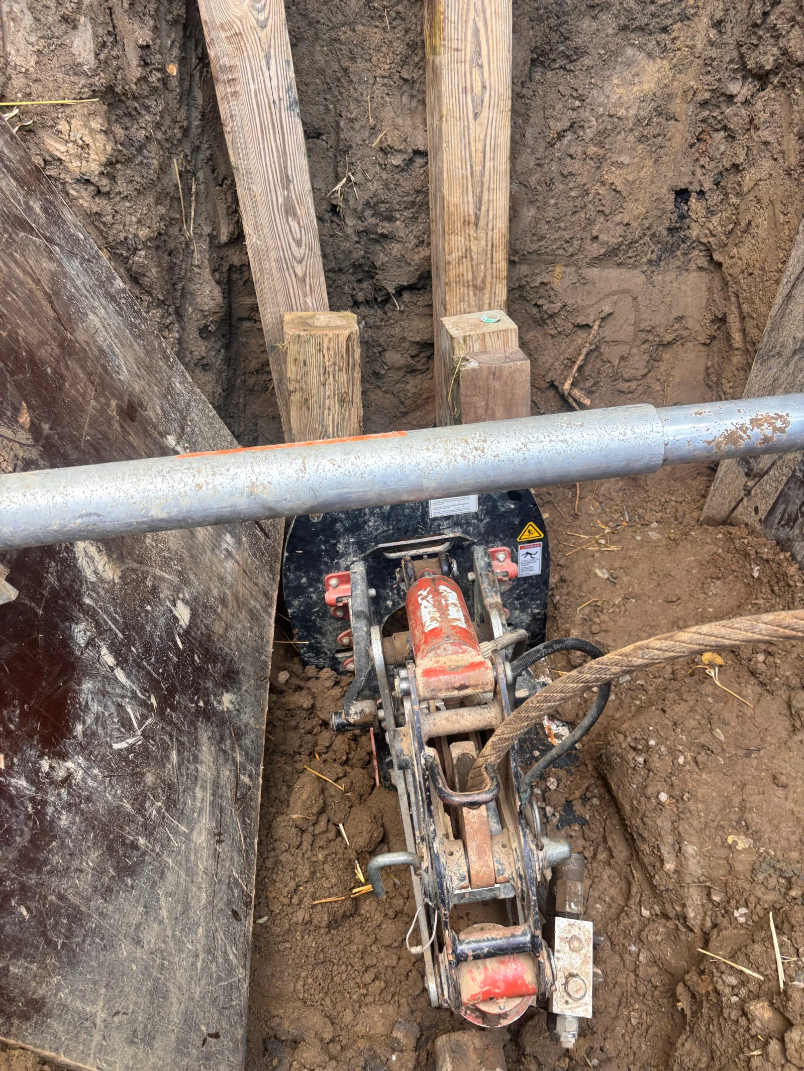 Pneumatic pipe bursting equipment with metal pipe in trench, wooden shoring supports visible
