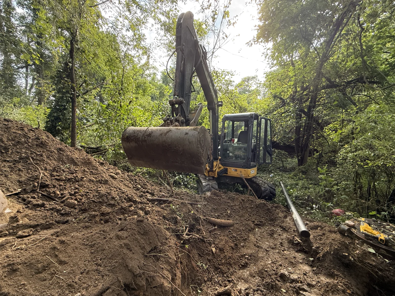 Excavator installing large drainage pipe in wooded area with minimal ground disturbance